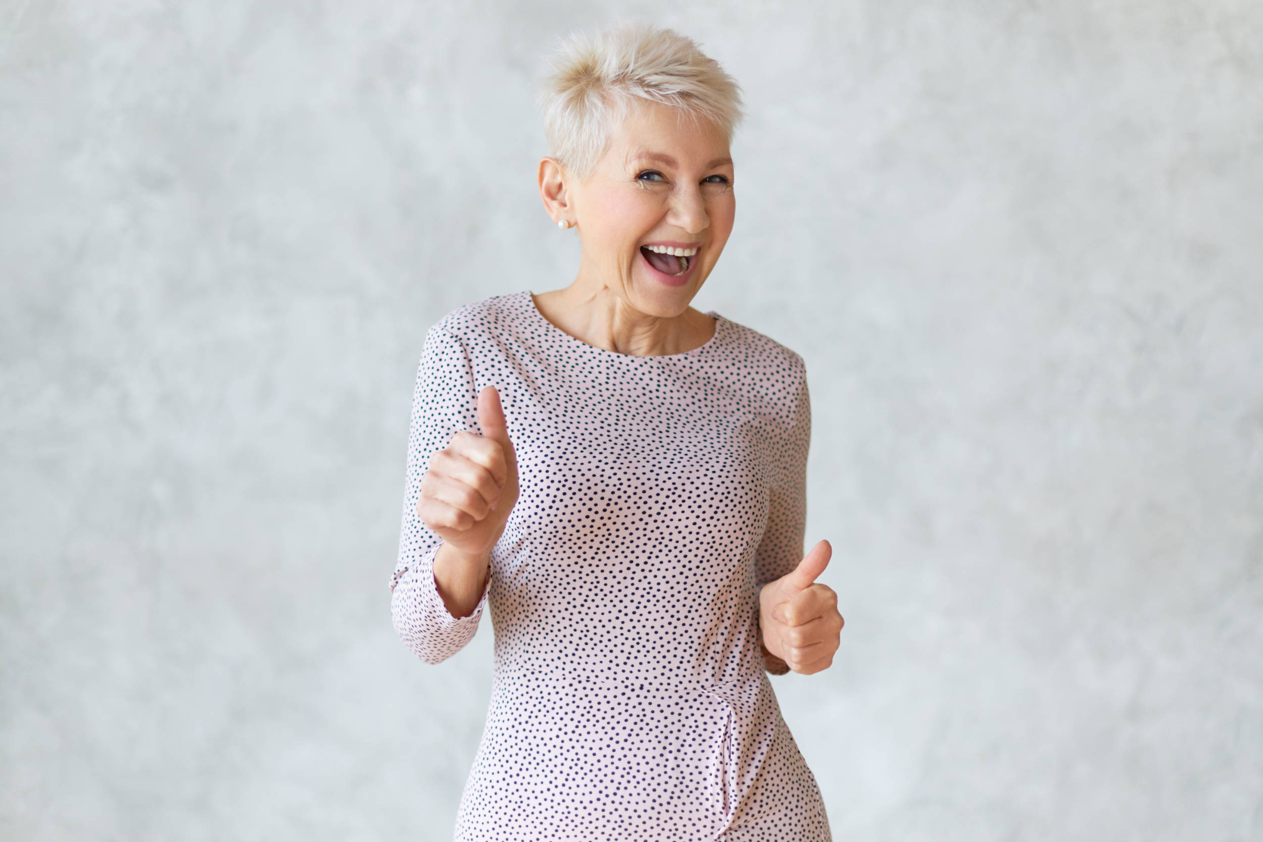 Funny cheerful middle aged blonde woman wearing elegant pencil dress dancing and showing thumbs up gesture as sign of approval, celebrating success or profitable deal, smiling broadly at camera